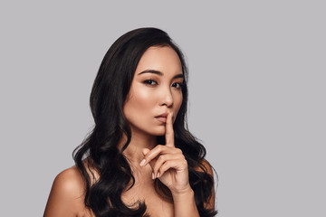 Naklejka premium Can you keep a secret? Beautiful young woman looking at camera and keeping finger on lips while standing against grey background