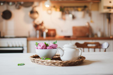 Obraz na płótnie Canvas White teapot and pink cupcakes are serving on straw wicker tray on table. Tea time, cozy home atmosphere hygge. Kitchen still life in warm light. Sweets with mint herbal leaves.