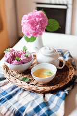 Obraz na płótnie Canvas Pink cupcakes, mug of green mint tea, white teapot and hydrangea flowers on kitchen table. Breakfast with healthy sweets is serving on straw tray. Tea time, cozy home atmosphere. Summer still life.