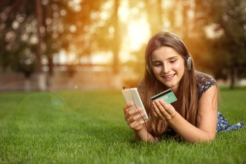 Obraz premium Portrait of a happy woman buying online with a smart phone outdoors lying down on green lawn headphones on head. Multicultural model, mixed race, asian russian girl. Copy space. People and technology.