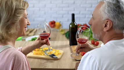 Loving aged couple holding glasses with red wine sitting home kitchen, cooking
