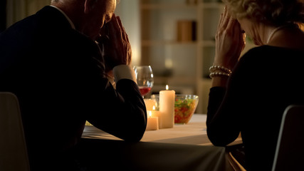 Aged religious husband and wife praying at table, joining hands, gratitude