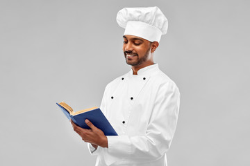cooking, profession and people concept - happy male indian chef in toque reading cookbook over grey...