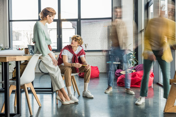focused female and male casual businesspeople sitting and having discussionin in loft office with colleagues in motion blur on background