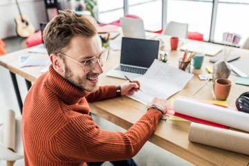 handsome smiling male architect sitting at desk and working on blueprints in loft office