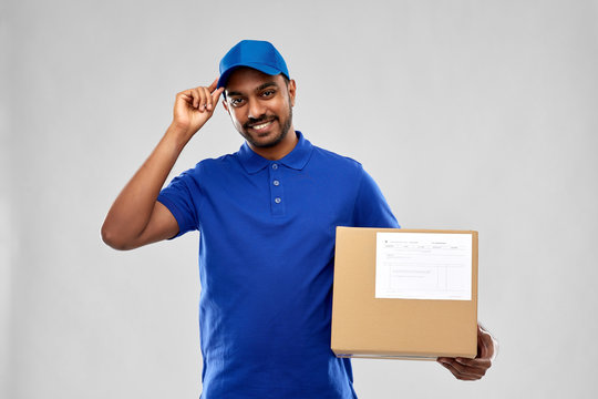 mail service and shipment concept - happy indian delivery man with parcel box in blue uniform over grey background