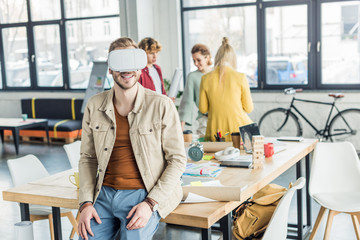 Naklejka premium smiling male designer having virtual reality experience in loft office with colleagues on background