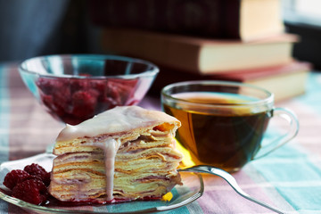 Homemade layer cake with raspberry vanilla custard dripping decorated with frozen berry near window sill with books on background, table setting: cups of tea, spoons, dessert , close up, copy space