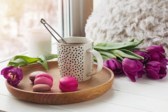 A cup of tea, sweet dessert macarons and purple tulips on a window sill.