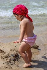 A cute and sweet little girl plays with water and sand on the seashore. Child with pink swimsuit and red bandana. Sea foam and waves.