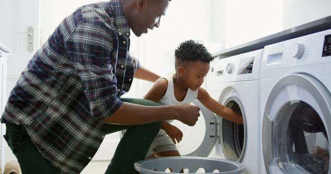 Side view of black father and son washing clothes in washing machine at comfortable home 4k