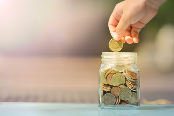 Kid's hand holding coin to put in glass piggy bank fulled of coins on blurred background with copy space. For business, teaching children, and saving concept.