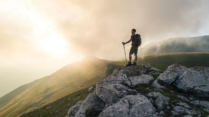 Young hiker man with backpack and walking poles, standing on peak of a mountain looking at sunset...