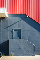 A pedestrian door in the black painted wall of a large warehouse, at the top of a small staircase, under a red corrugated metal siding with a security camera.