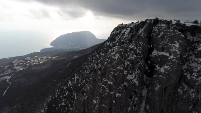 The slope of the high rock massif covered with pine trees in misty clouds. Shot. Aerial for the forested mountain slope in heavy, grey clouds and coastline with smal town.