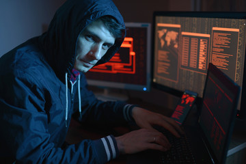 Male in the hood looking in the camera. Hacker attacks and online fraud in the dark on the screen...