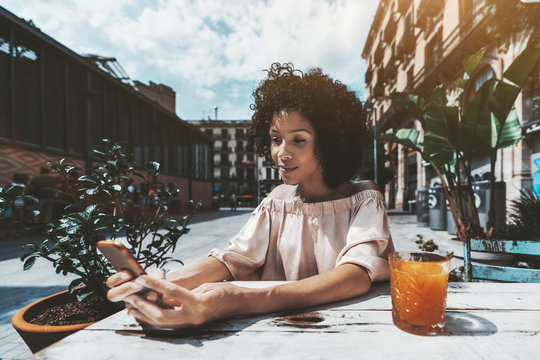 A pensive curly-hair African-American girl in a street bar is reading the feed of her friends in social networks using the smartphone, mango juice in the glass near her, sunny summer day in Barcelona