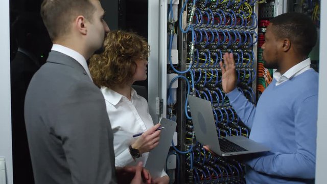 African american IT engineer holding laptop and telling about server rack to male and female Caucasian colleagues while working together in data center
