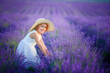 Cute curly young girl standing on a lavender field in white dress and hat with cute face and nice hair with lavender bouquet and smiling.