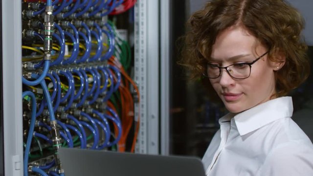 Close up view of young female IT engineer in glasses standing beside opened server rack in data center and working on laptop computer while testing equipment