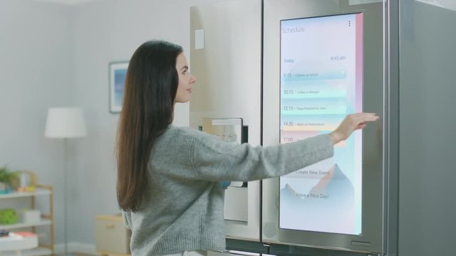 Beautiful Young Girl Walks Over to a Refrigerator While Drinking Her Morning Coffee. She is Checking the Weather Forecast and a To Do List on a Smart Fridge at Home. Kitchen is Bright and Cozy.