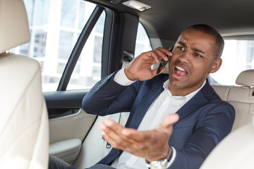 Young businessman sitting on back seat in car talking on smartphone shouting at driver irritated