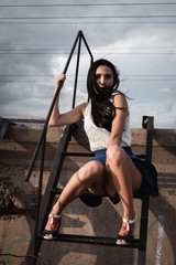 Girl in skirt sitting on the iron stairs bent legs. The wind is blowing through her hair. Behind...