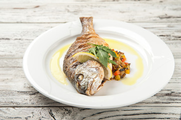 Dorada stuffed with vegetables. White plate with grilled dorada fish on a wooden table close up and copy space