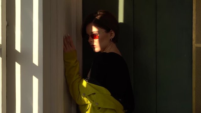 Brunette girl with short hair and sunglasses posing against the wall. She leans her hand against the wall.First she wears a light green jacket, then she takes it off