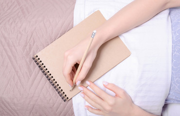 Woman in the white bed in hand notebook pencil brown blanket, top view