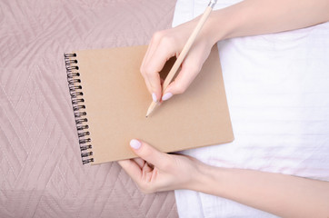 Woman in the white bed in hand notebook pencil brown blanket, top view