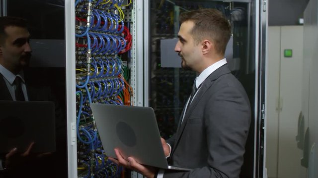 Male IT technician in formal suit standing beside server rack in data center, inspecting wires and typing on laptop