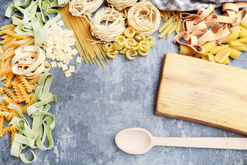 Wooden cutting board and mixed types and shapes of italian pasta on grey stone background