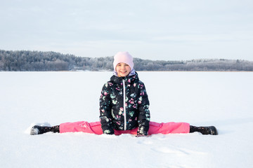 Fototapeta na wymiar Arctic outdoor winter portrait of a young child girl in winter clothing sitting in split in the snow against frozen landscape background.