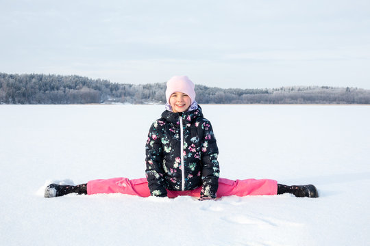 Arctic outdoor winter portrait of a young child girl in winter clothing sitting in split in the snow against frozen landscape background.