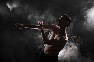 Fototapeta na wymiar A young male ballet dancer with black leggings and a naked torso performs dance moves against a gray grunge background, with a light of lights and smoke.