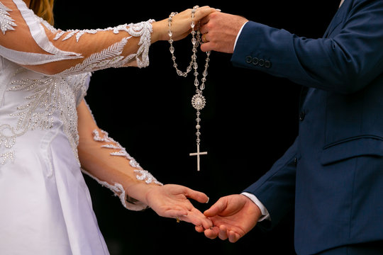 M&atilde;os de uma mulher e um homem, noivos, segurando um ter&ccedil;o com crucifixo em fundo preto