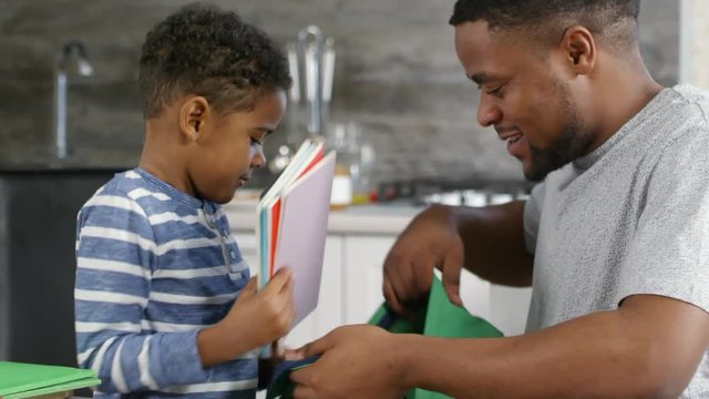 Close up of cute African boy and his father putting pencils, lunchbox and notebooks into school backpack together
