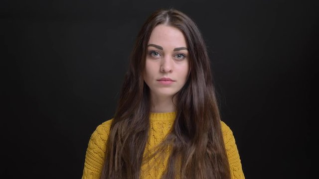 Portrait of young long-haired brunette caucasian girl in yellow sweater watching seriously into camera on black background.