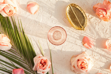 Fototapeta na wymiar Top view of stylish composition with pink peony roses flowers, tropical palm leaf, golden plate and glass on pastel textured background. Flat lay of home desk with shadows and sunlight.