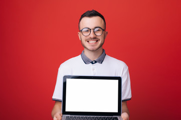 Young hispanic man wearing white tshirt and black glasses holding open laptop in his hands and smiling isolated on red background