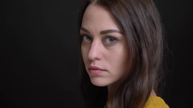 Close-up portrait of long-haired brunette girl in yellow sweater turns to camera and watches seriously on black background.