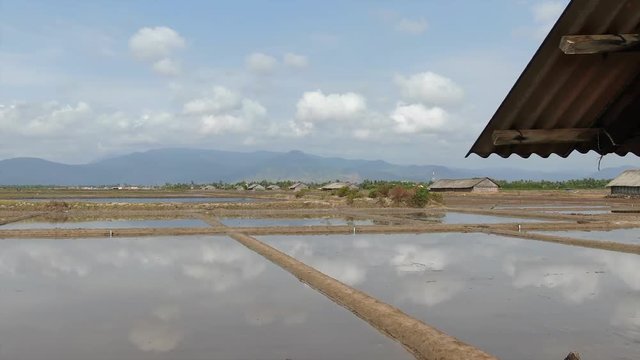 Aerial low view fly slowly towards flooded fields in Asia, with outline of wooden shack on the right. Settle on view of fields reflecting the sky in the water.