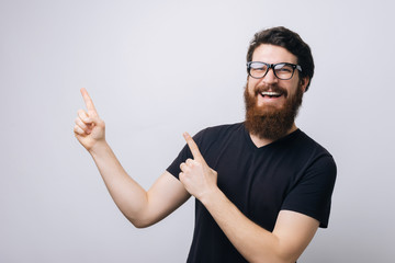 Young bearded man in black tshirt and glasses, smiling and pointing at copyspace while looking at...