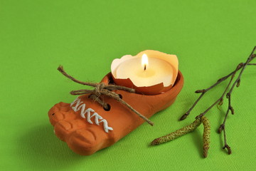 Lighted wax candle in eggshell in decorative clay boot with jute cord and birch twigs with earrings on bright green background. Symbolic concept - revival of new life.