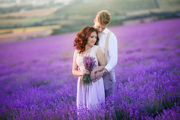 Couple is hugging and standing on lilac lavender flowers field at blurred background of mountain, nature after wedding ceremony. Bride is dressed in white wedding dress. Bride and groom are married.