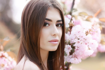 Fototapeta premium The beautiful girl with a romantic hairstyle and a professional make-up enjoys a smell of pink colors in a garden. The girl dreams. A portrait of the beautiful girl model in the spring in the park.
