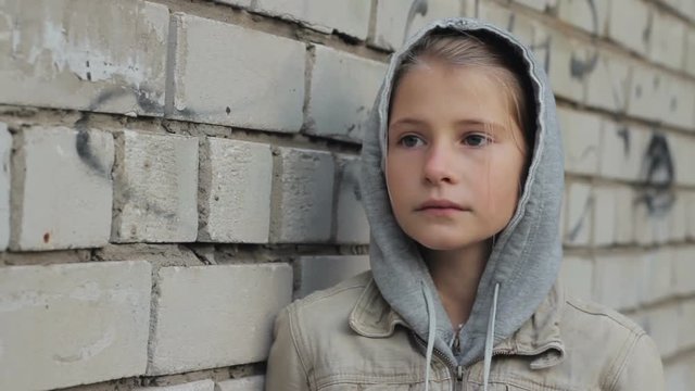 Sad depressed young girl portrait against grey brick wall. Upset lonely child wearing hooded jacket raises and lowers her head, sighs outdoors, steadicam shot.