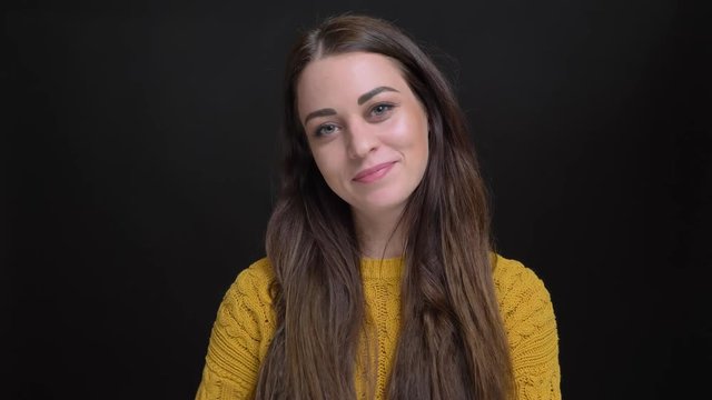 Portrait of long-haired brunette girl in yellow sweater humbly smiling into camera and fixing hair on black background.