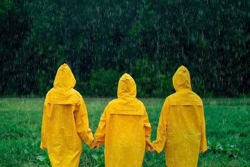 Three girls in yellow raincoats holding hands, standing in middle during rainfall in front of deciduous forest. Travel lifestyle concept vacations outdoor. Children looks at forest in distance